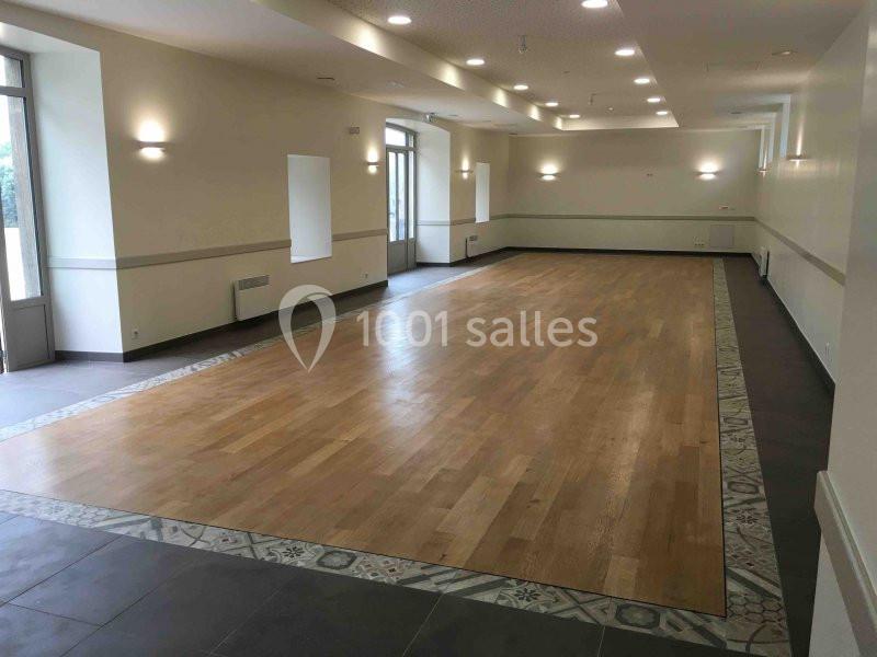 Salle vide avec parquet central entouré de carrelage, murs clairs, éclairage mural et grandes fenêtres.