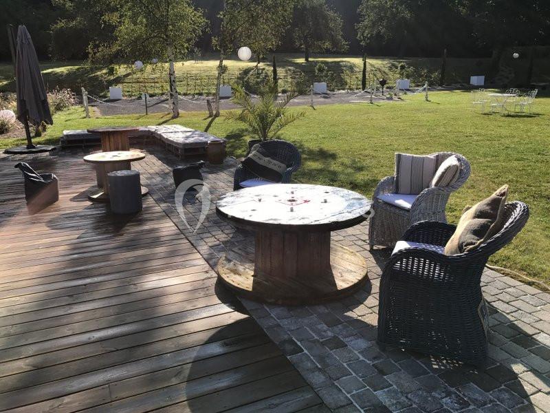 Terrasse en bois avec mobilier extérieur composé de fauteuils en osier et tables rondes, entourée de verdure.
