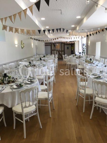 Salle de réception décorée avec des guirlandes et des tables rondes dressées avec nappes blanches et chaises blanches.
