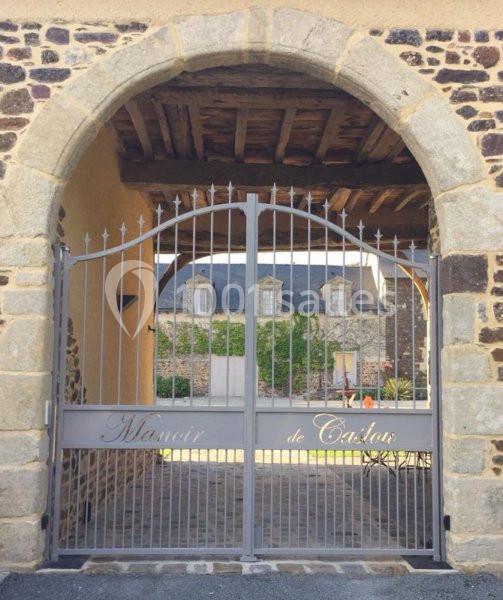 Portail en fer forgé sous une arche en pierre, donnant sur une cour pavée et un bâtiment en arrière-plan.