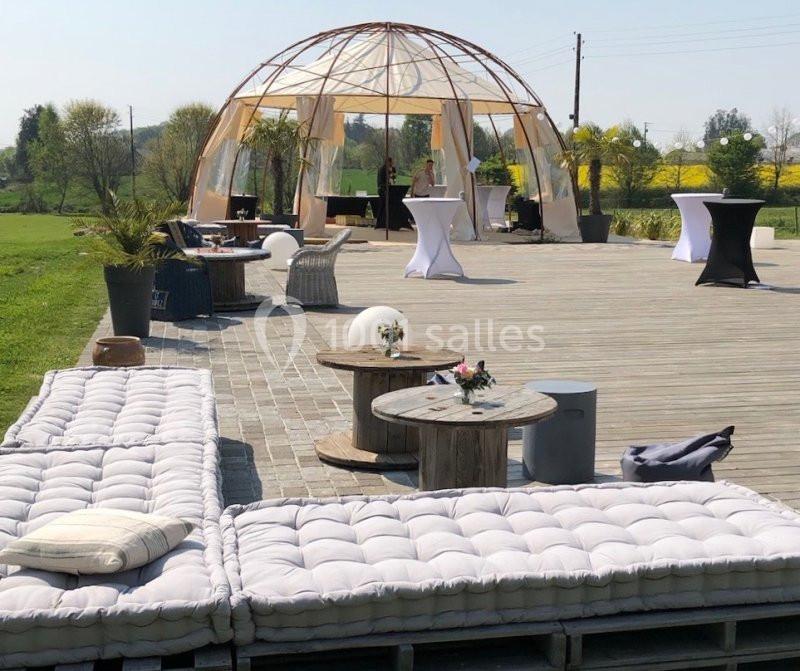 Terrasse en bois aménagée avec coussins, tables basses et une structure en toile dans un cadre champêtre.