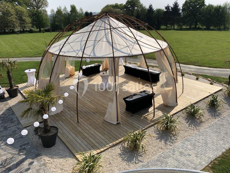 Structure en toile beige sur une terrasse en bois, entourée de plantes et de lanternes, dans un espace vert.