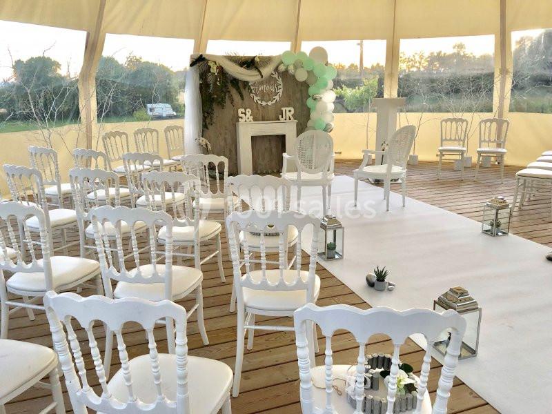 Chaises blanches disposées pour une cérémonie dans une tente décorée avec des ballons, lanternes et un tapis blanc.