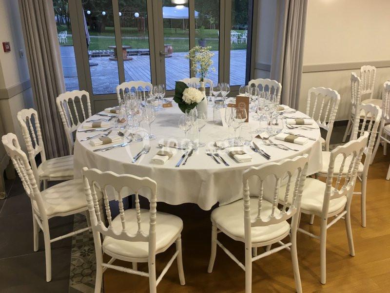Table ronde dressée avec nappes blanches, couverts, verres et centre de table floral, entourée de chaises blanches.