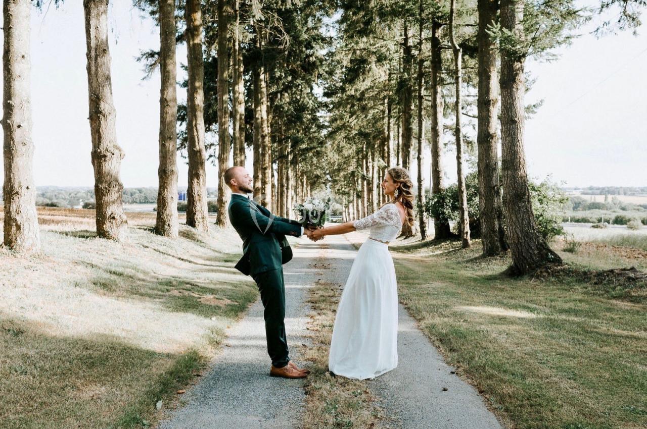 Un couple en tenue de mariage se tient par les mains sur une allée bordée d'arbres par une journée ensoleillée.
