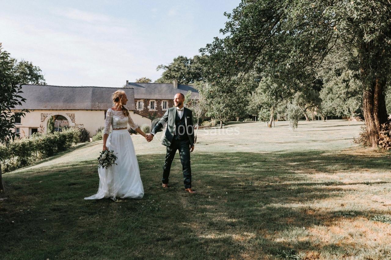 Un couple en tenue de mariage marche main dans la main dans un jardin verdoyant près d'un bâtiment en pierre.