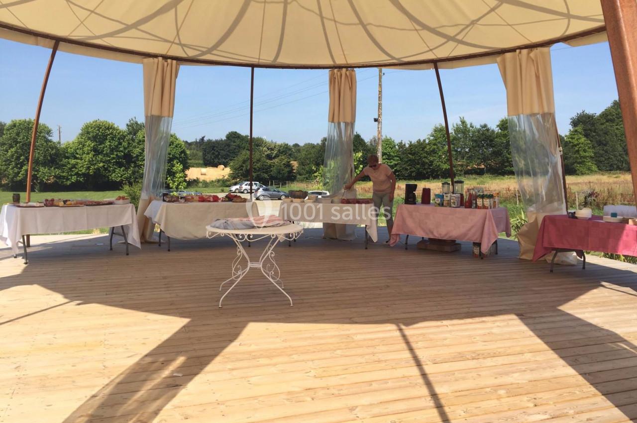 Tables dressées sous une grande tente ouverte, avec nappes blanches et roses, dans un cadre extérieur ensoleillé.