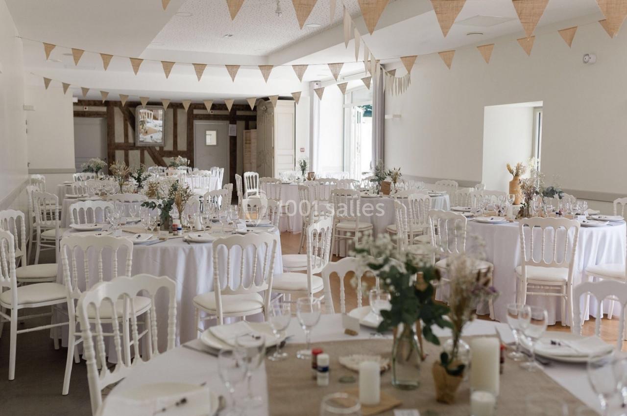 Salle de réception décorée pour un événement, avec des tables rondes dressées et des guirlandes en tissu suspendues.