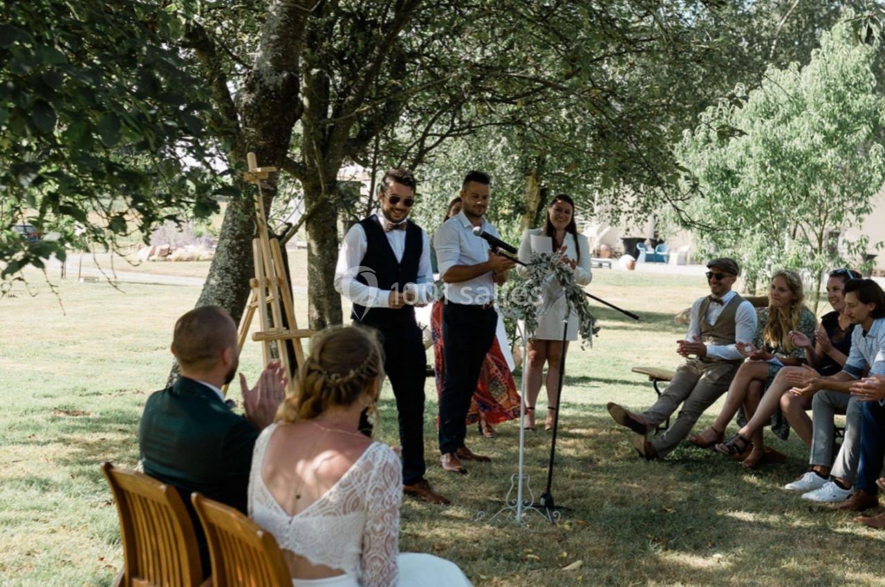Un couple assis écoute un discours lors d'une cérémonie en plein air sous des arbres, entouré d'invités.