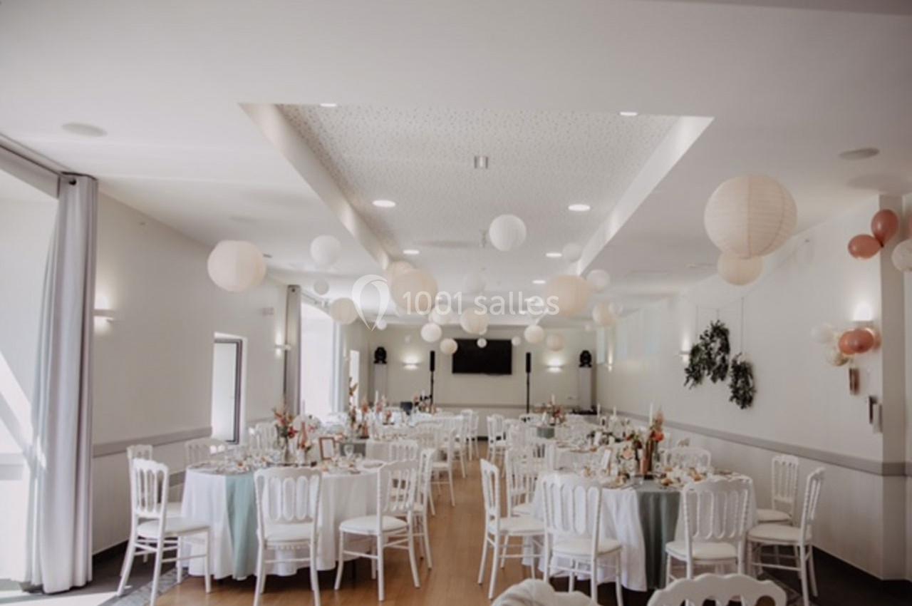 Salle de réception lumineuse décorée de lanternes suspendues, avec des tables rondes dressées et des chaises blanches.