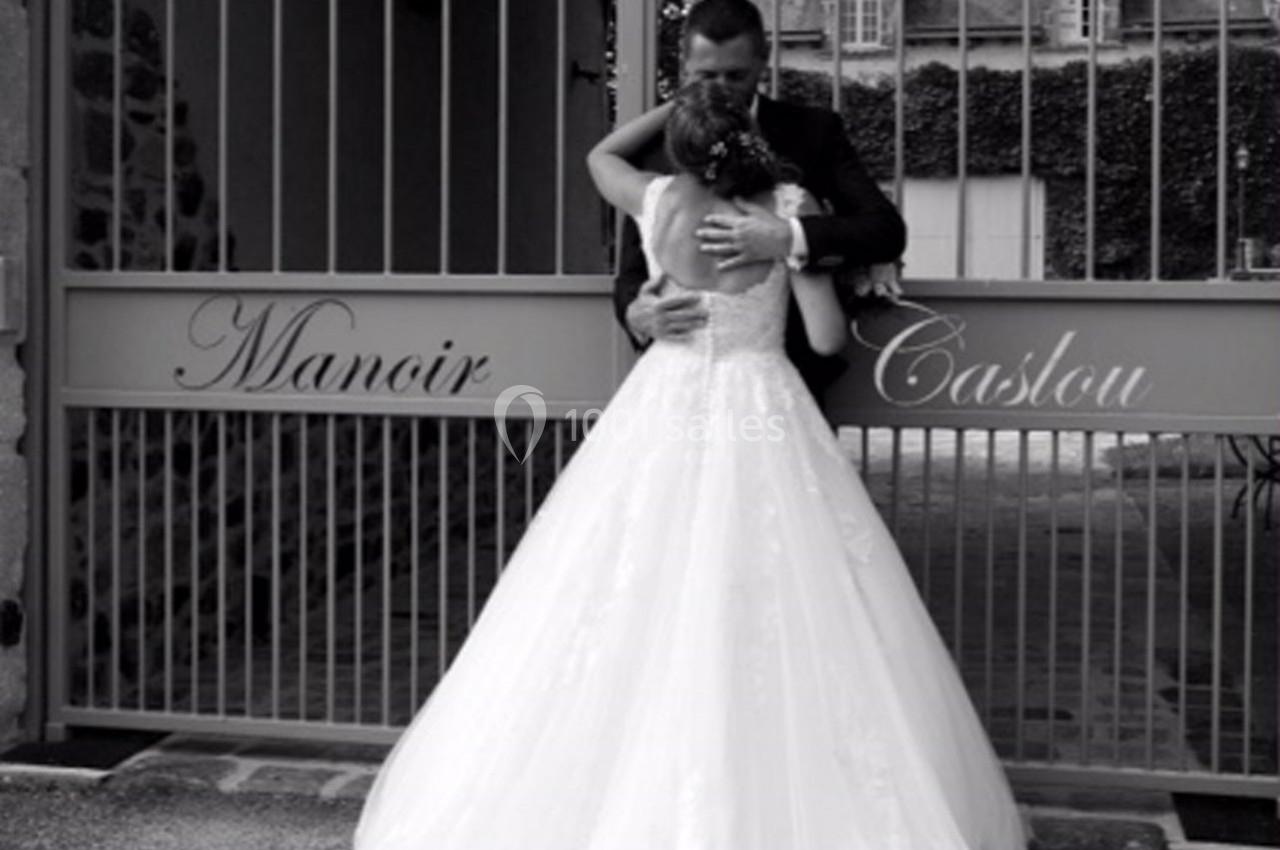Un couple en tenue de mariage s'enlace devant une grille portant l'inscription ’Manoir Caslou’.