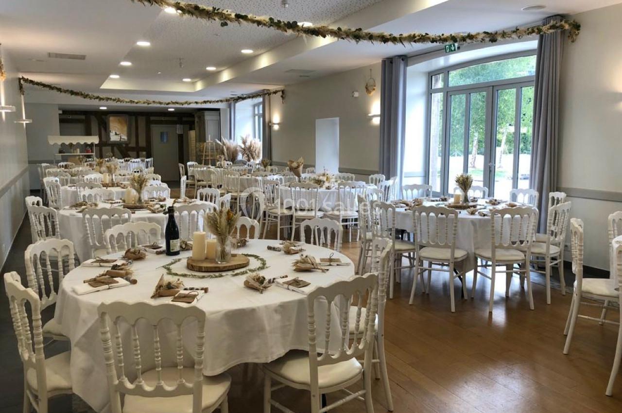 Salle de réception décorée avec des tables rondes, nappes blanches, chaises en bois et ornements champêtres.
