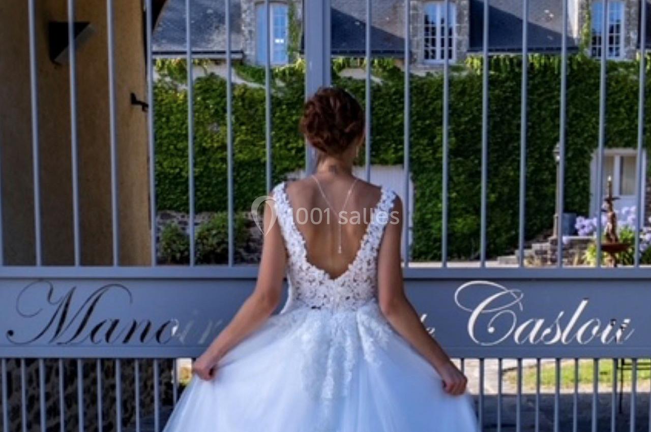 Une femme en robe de mariée blanche vue de dos, devant une grille métallique ornée d'une inscription.