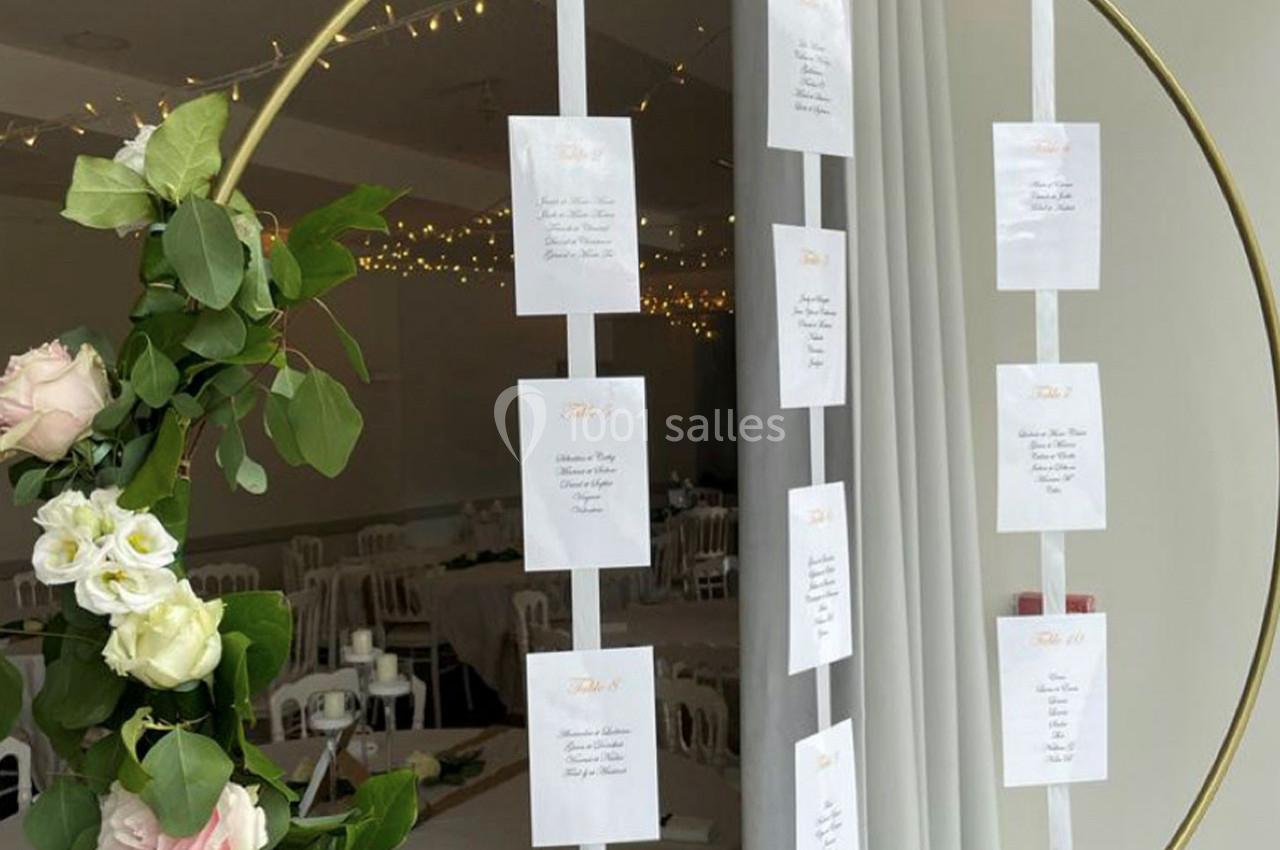 Cercle doré décoré de fleurs supportant des cartes de placement suspendues, dans une salle de réception lumineuse.