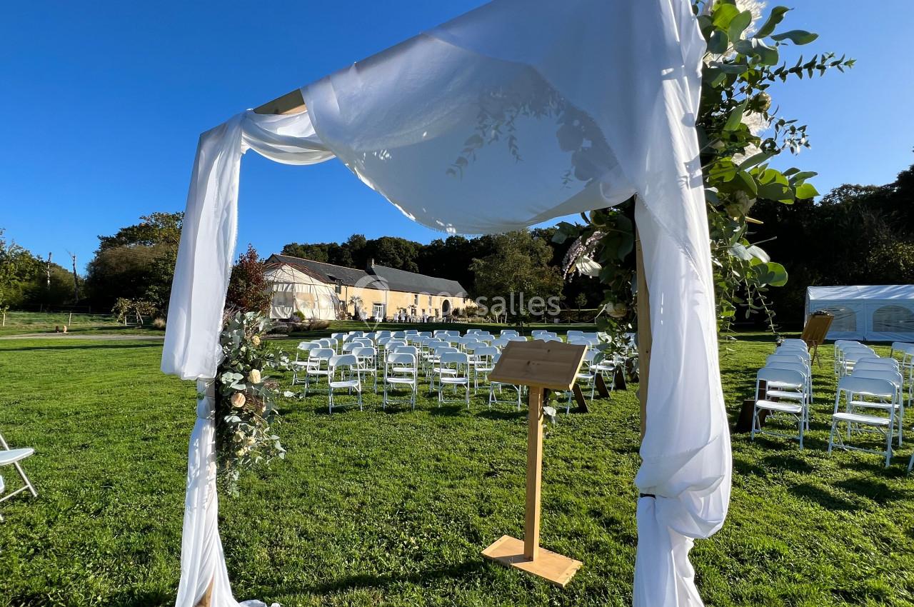Arche de cérémonie décorée de tissu blanc et de feuillage, installée dans un jardin avec des chaises alignées en arrière…
