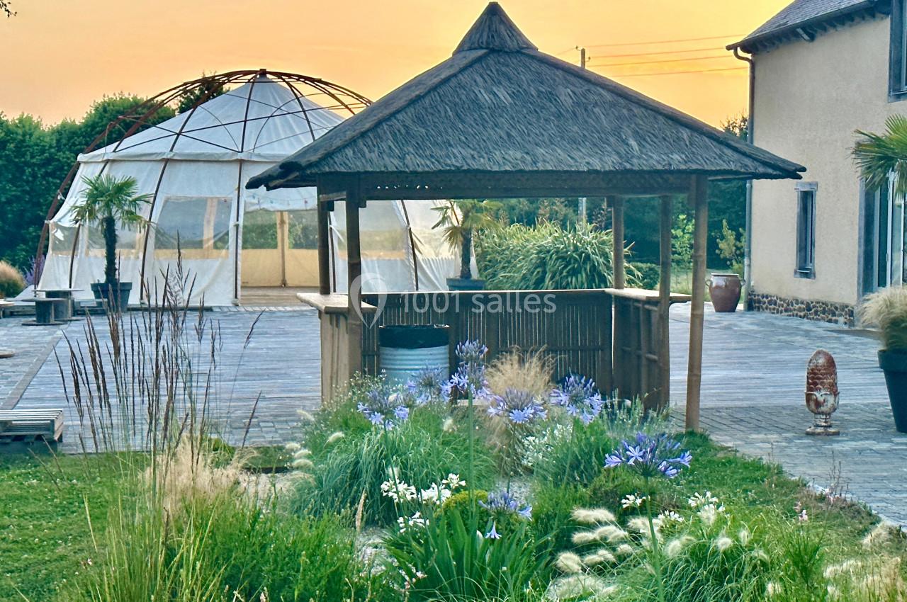 Kiosque en bois avec toit en chaume entouré de fleurs, près d'une terrasse et d'une structure en dôme, au coucher du soleil.