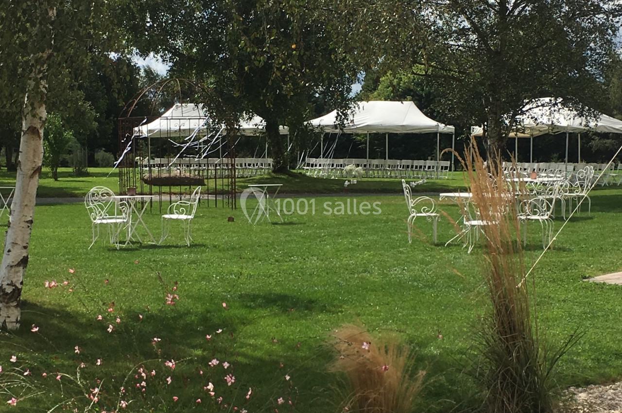 Chaises et tables blanches disposées sur une pelouse entourée d'arbres, avec des tentes blanches en arrière-plan.