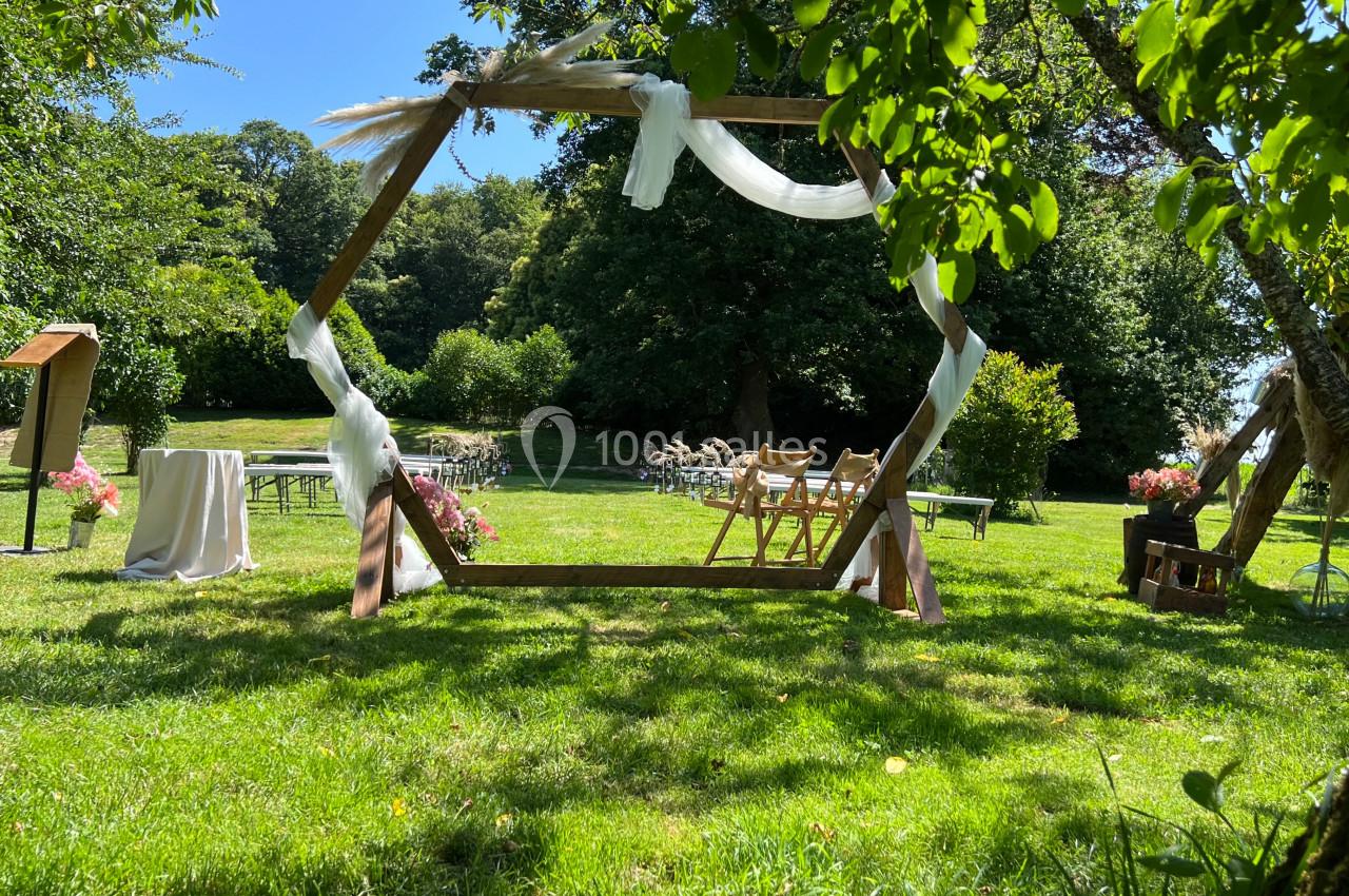 Cérémonie en plein air avec arche décorée de tissu blanc, chaises en bois et verdure environnante.