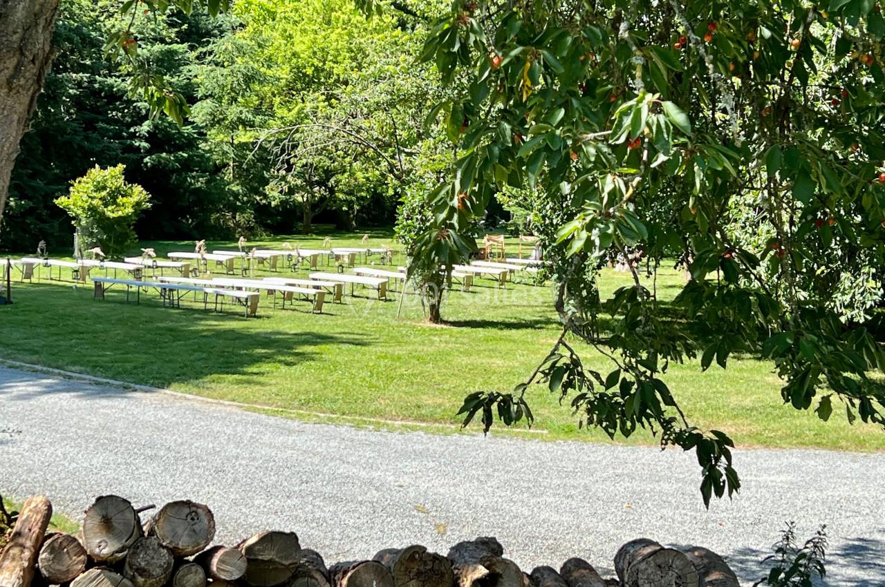 Tables et bancs disposés sur une pelouse verdoyante, entourés d'arbres, avec un chemin et des bûches au premier plan.