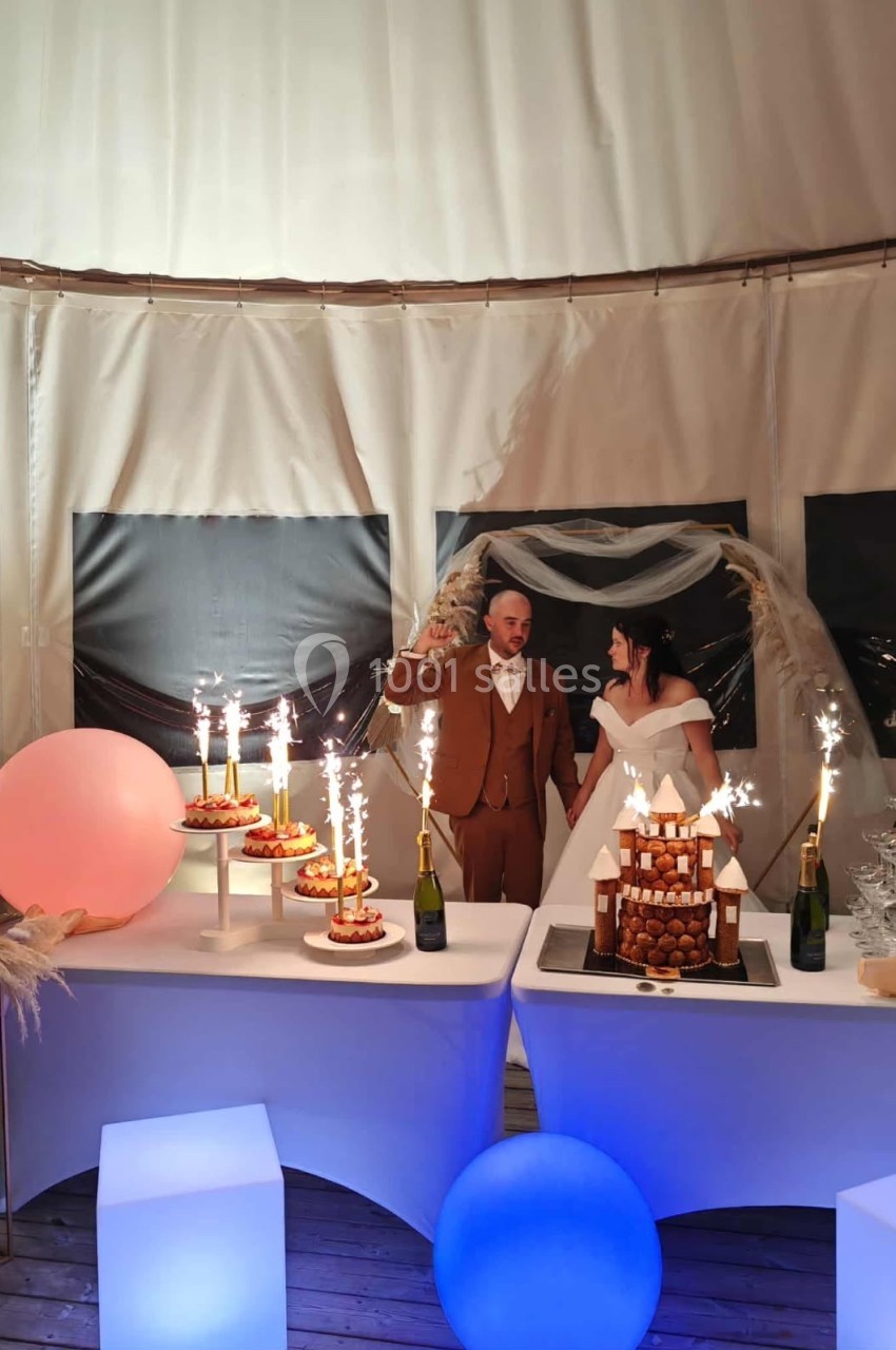 Un couple en tenue de mariage trinque devant une table décorée avec des desserts et des fontaines lumineuses.