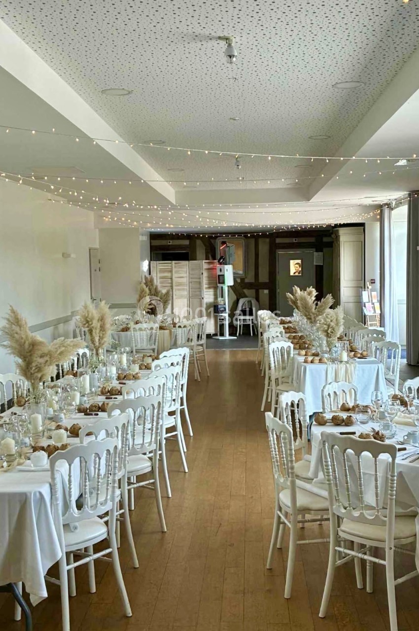 Salle de réception décorée avec des tables rondes, nappes blanches, chaises blanches et ornements de fleurs séchées.