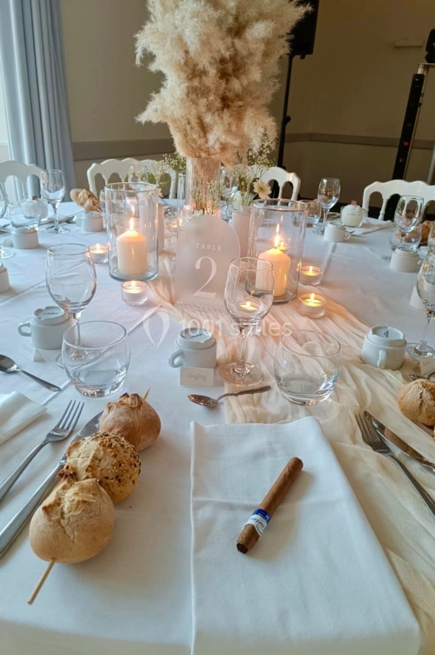 Table dressée pour un repas, avec bougies, fleurs séchées, vaisselle élégante et petits pains disposés autour.