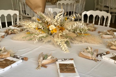 Centre de table floral avec fleurs séchées et pampas, entouré de couverts et cadeaux emballés sur une nappe blanche.