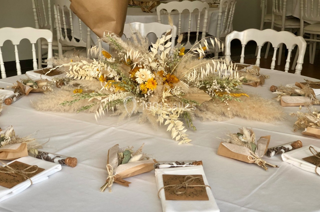 Centre de table floral avec fleurs séchées et pampas, entouré de couverts et cadeaux emballés sur une nappe blanche.