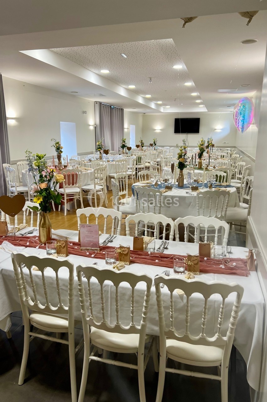 Salle de réception décorée avec des tables dressées, nappes blanches, chemins de table roses et bouquets de fleurs.