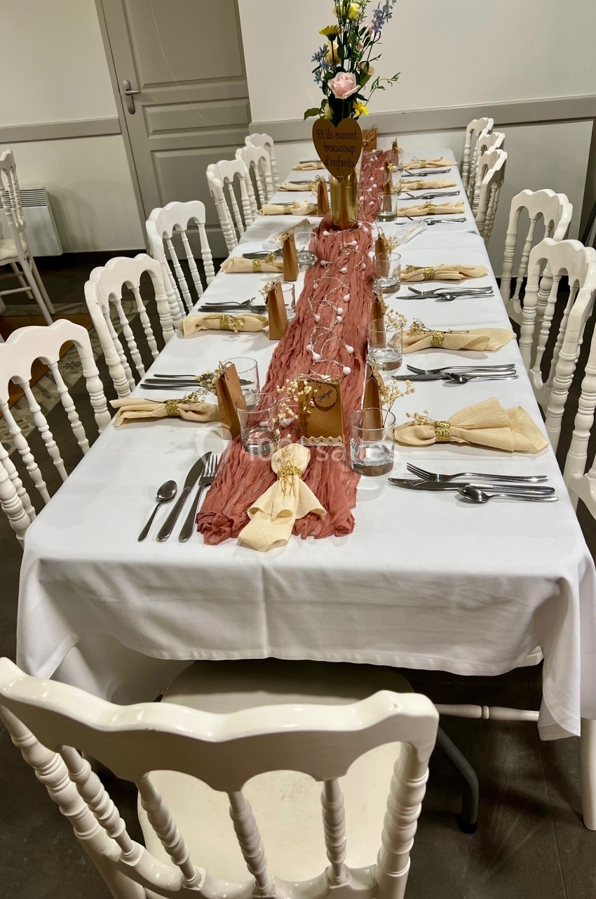 Table décorée pour un repas, avec nappe blanche, chemin de table rose, vaisselle, serviettes pliées et chaises blanches.