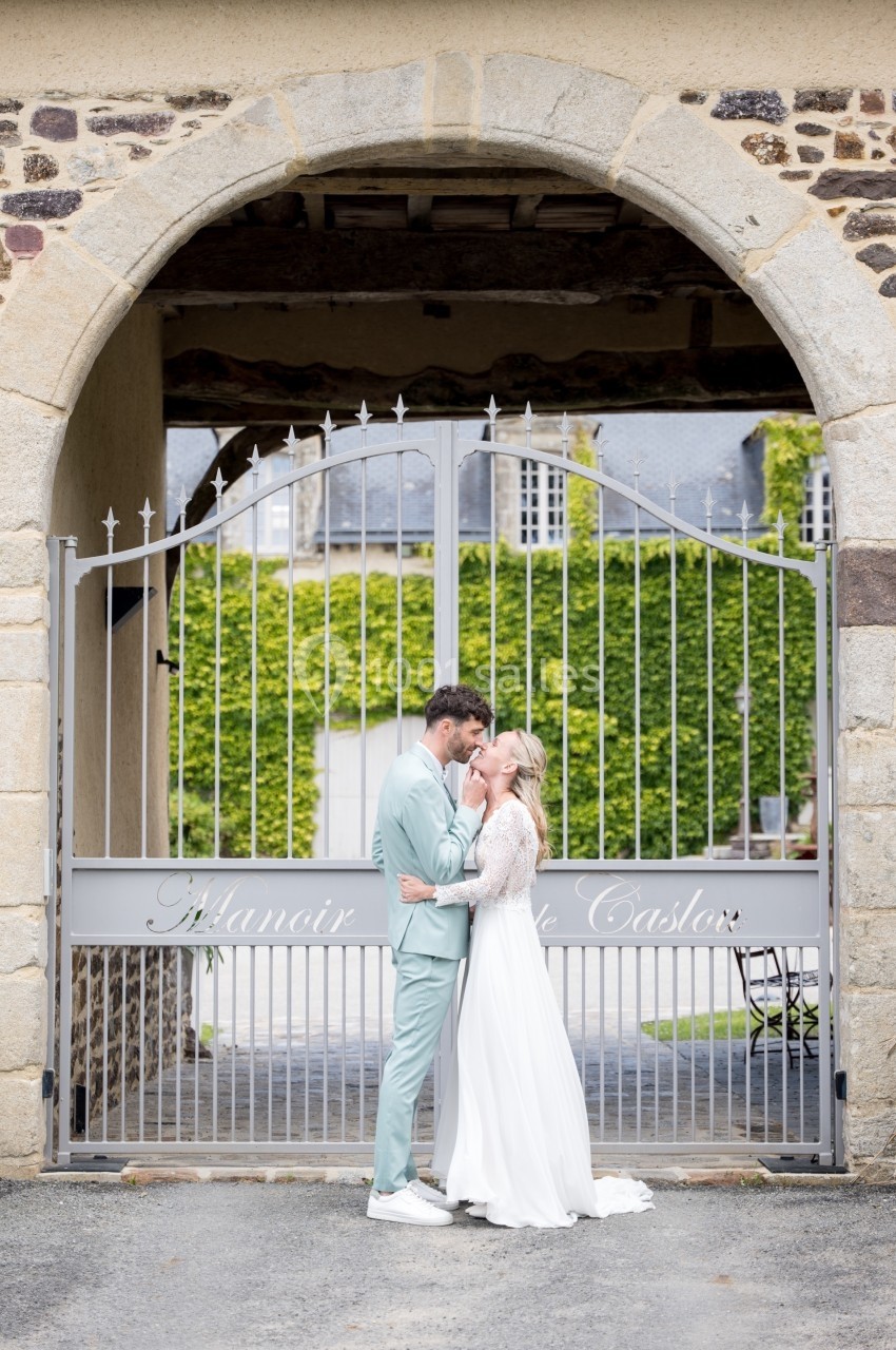 Un couple en tenue de mariage s'embrasse devant une grille en fer forgé sous une arche en pierre.