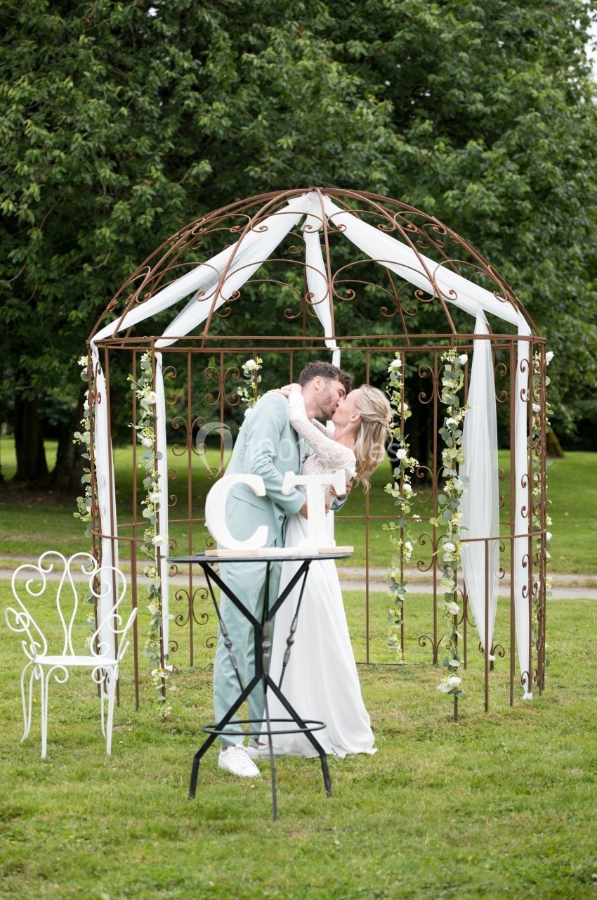 Un couple en tenue de mariage s'embrasse sous une pergola décorée de fleurs dans un jardin verdoyant.