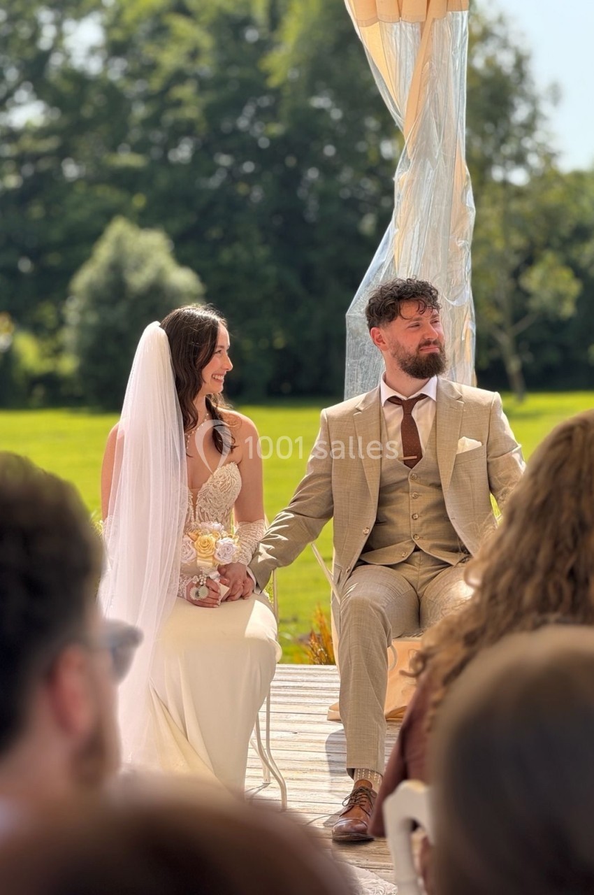 Un couple assis sous une pergola lors d'une cérémonie de mariage en extérieur, entouré d'invités.