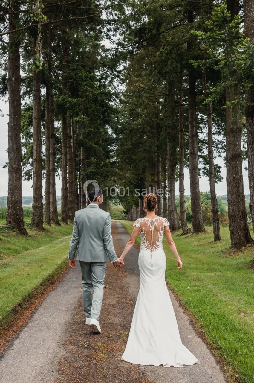 Un couple en tenue de mariage marche main dans la main sur une allée bordée d'arbres.