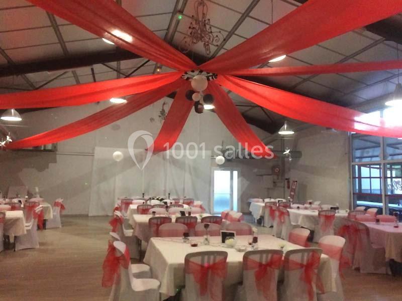 Salle de réception décorée avec des nappes blanches, des chaises ornées de rubans rouges et des tentures rouges au plafond.