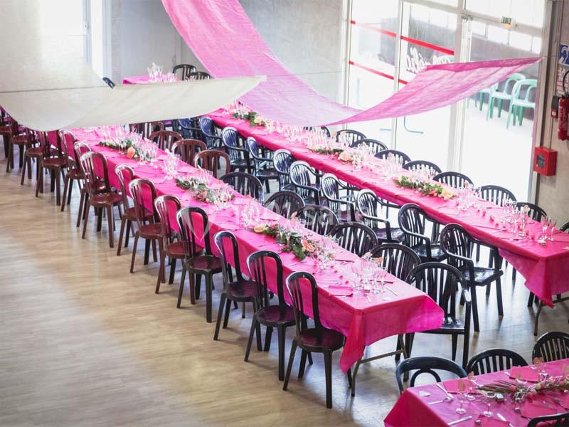 Salle décorée avec des tables longues recouvertes de nappes roses, disposées pour un événement festif.