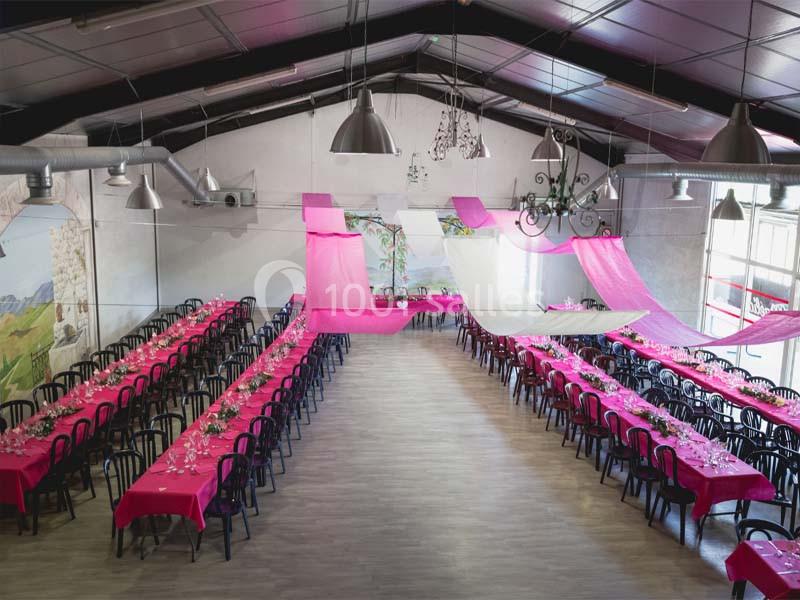 Salle de réception décorée avec des tables longues couvertes de nappes roses et des voilages suspendus au plafond.
