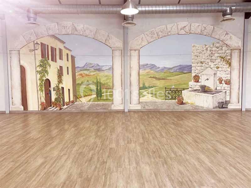 Fresque murale représentant un paysage rural méditerranéen avec maison, colline et fontaine, dans une salle au sol en bois.