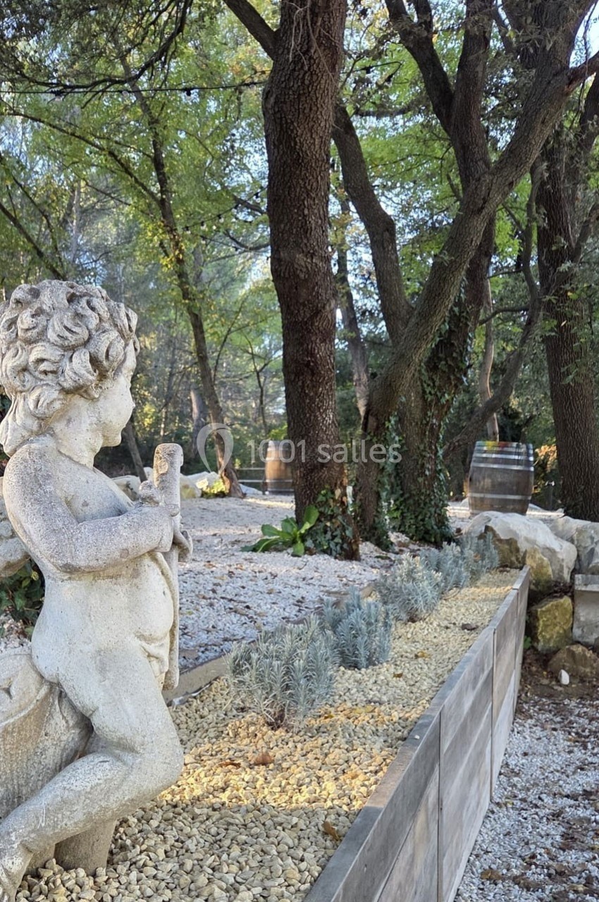Statue en pierre d'un enfant tenant un objet, entourée de végétation, avec des arbres et des tonneaux en arrière-plan.