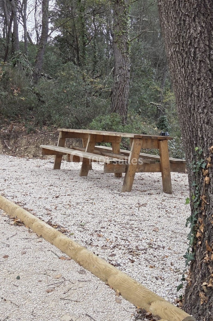 Table de pique-nique en bois sur un sol de gravier, entourée d'arbres dans un cadre naturel.