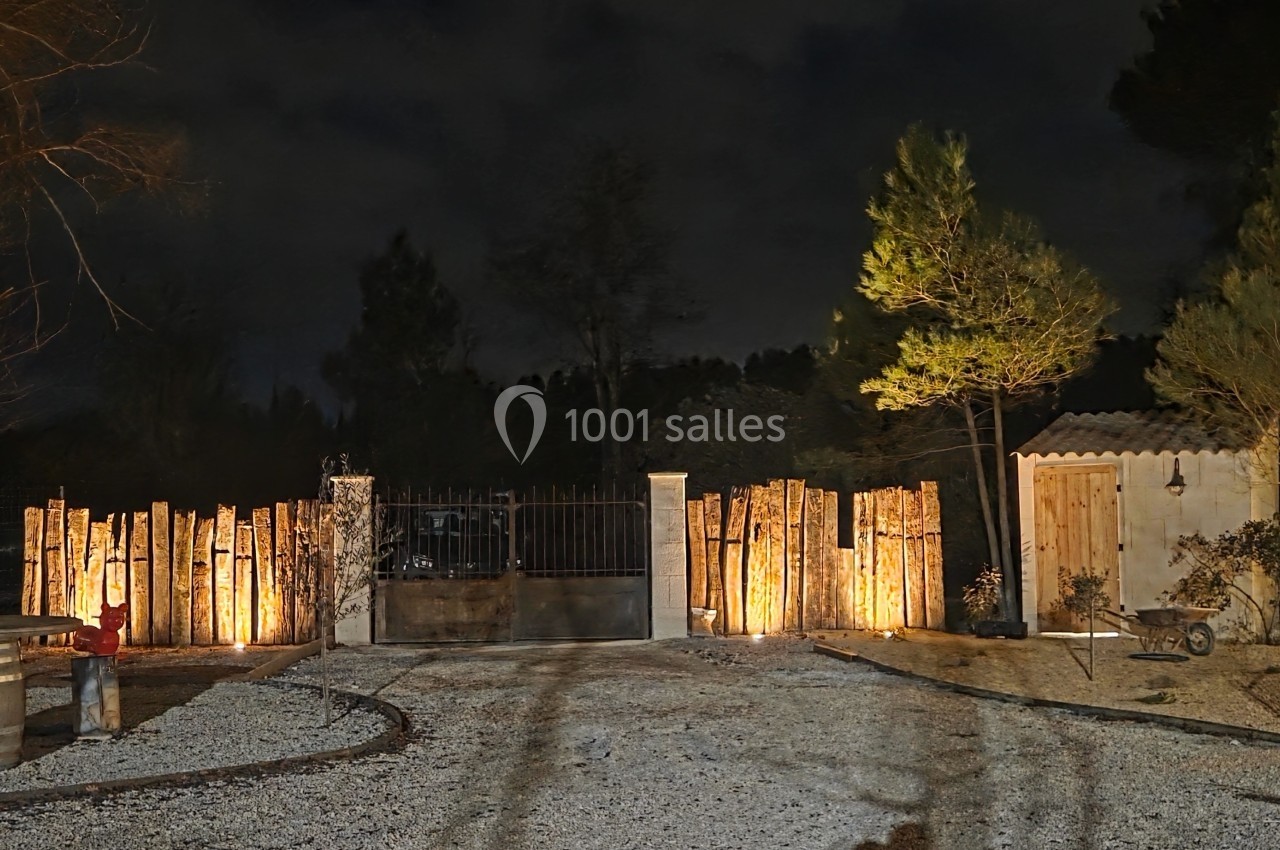 Entrée d'une propriété avec portail en métal, clôture en bois éclairée, et cabanon en arrière-plan, de nuit.