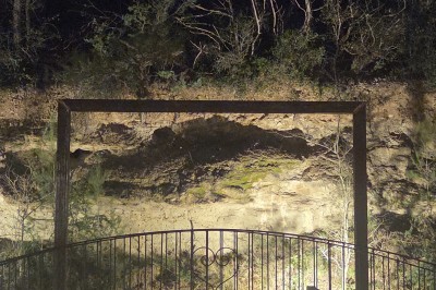 Clôture métallique devant un mur de pierre éclairé, entouré de végétation dans une ambiance nocturne.