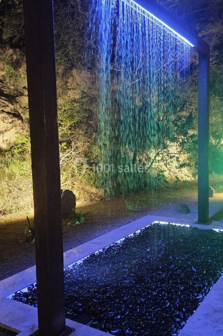 Location salle Loriol-du-Comtat (Vaucluse) - La Maelia #30 Cascade lumineuse artificielle avec éclairage bleu, tombant dans un bassin entouré de pierres, en extérieur nocturne.
