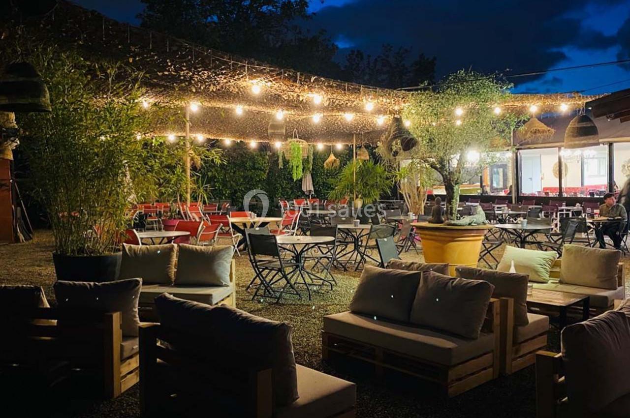 Terrasse extérieure éclairée en soirée avec des guirlandes lumineuses, des tables, des chaises et des plantes.