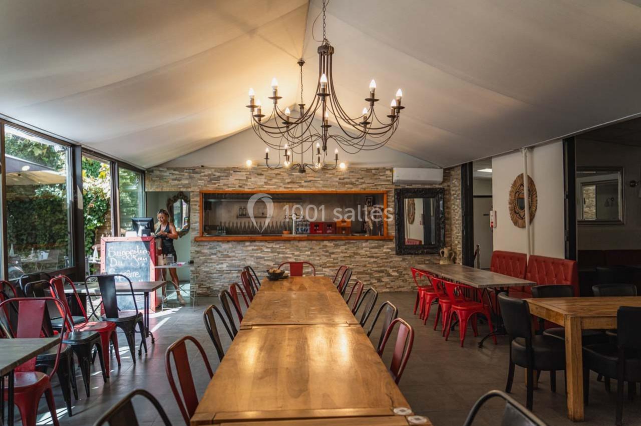 Salle de restaurant lumineuse avec de longues tables en bois, des chaises colorées et un lustre central.