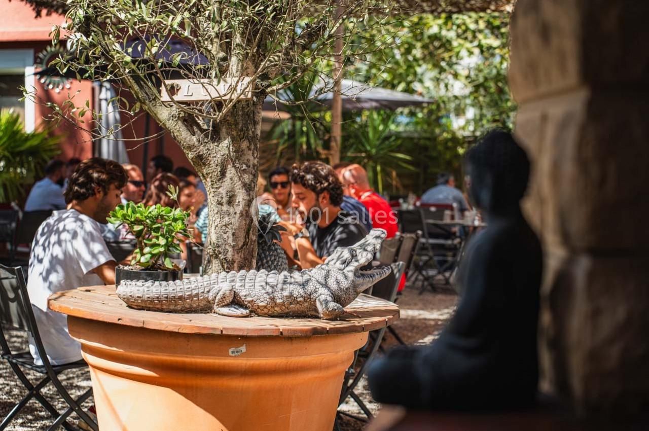 Un crocodile décoratif en céramique posé sur un pot en terrasse ombragée avec des personnes attablées en arrière-plan.
