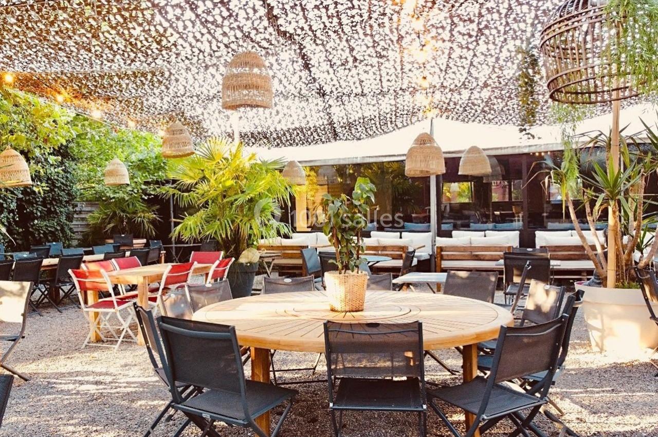Terrasse extérieure aménagée avec tables rondes, chaises noires, végétation et luminaires suspendus sous une toile ajourée.
