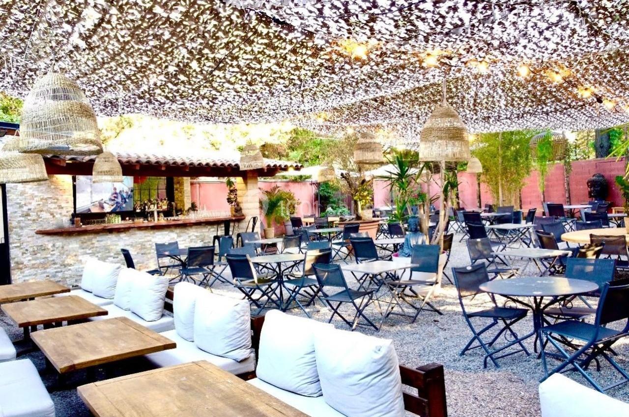 Terrasse extérieure avec tables, chaises pliantes, coussins blancs, bar en pierre et décorations lumineuses suspendues.