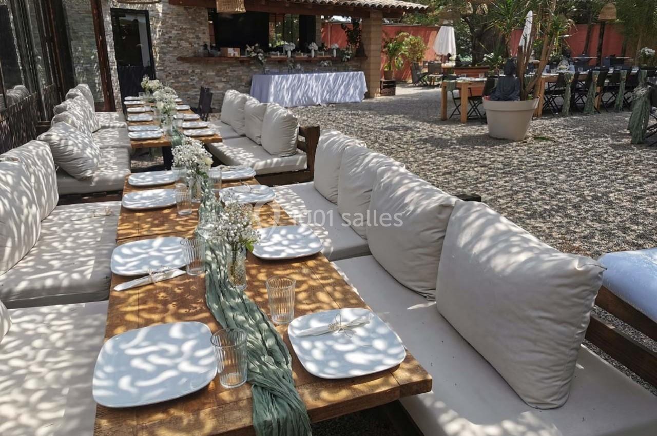 Tables en bois dressées avec des assiettes blanches et des décorations florales, dans une cour extérieure ombragée.
