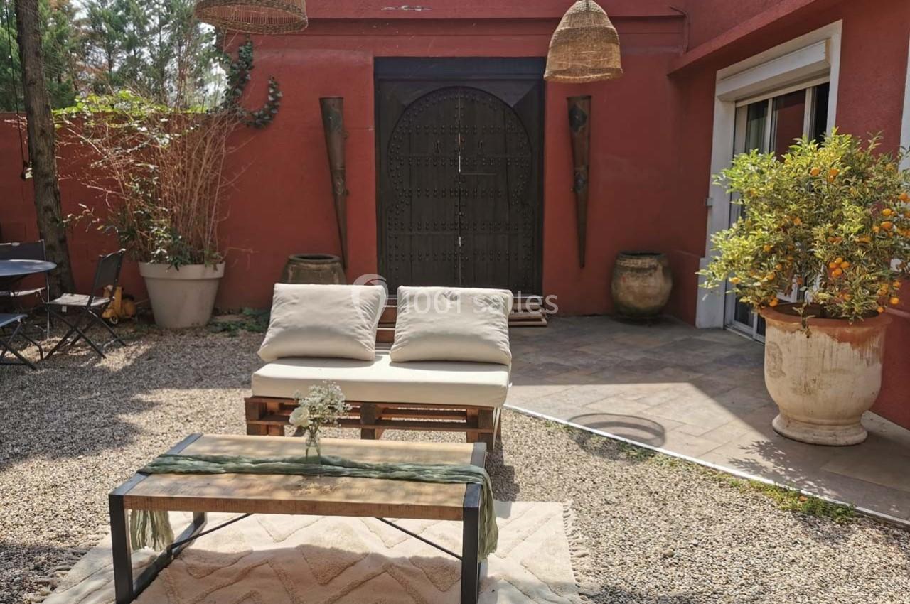 Salon de jardin extérieur avec canapé en palettes, table basse et plantes, devant une façade rouge avec porte en bois.